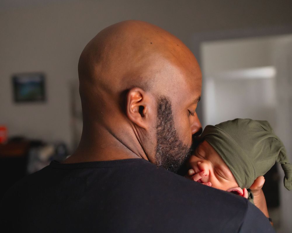 A tender moment showing nurturing care between a person wearing black and a baby in an olive-colored hat.