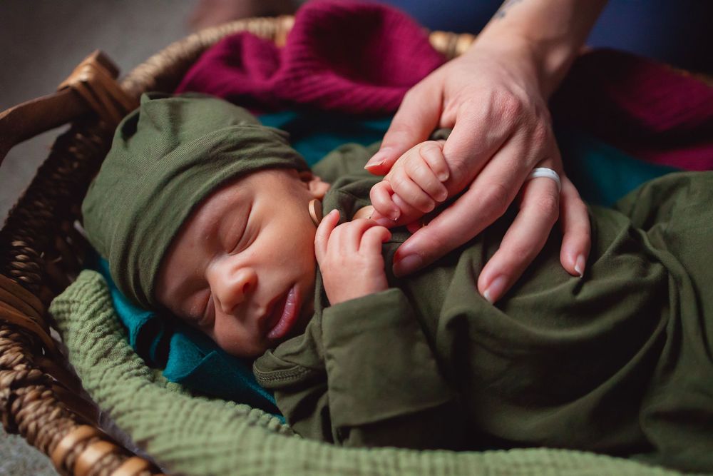 A sleeping baby dressed in olive green clothing rests peacefully in a woven basket with colorful bedding beneath.