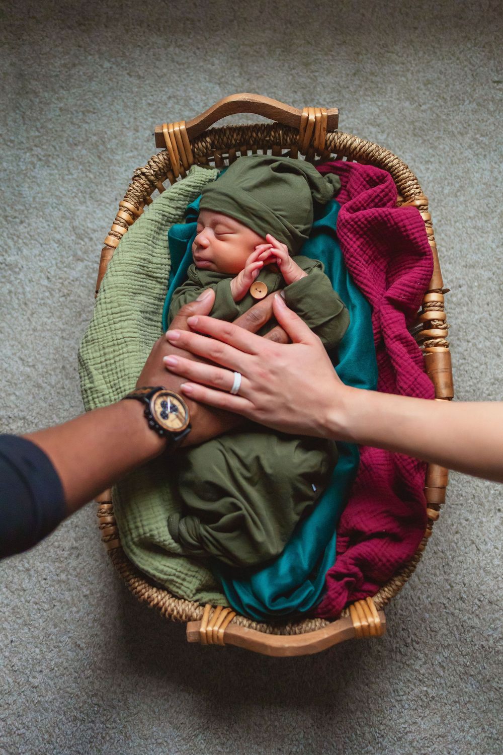 A sleeping infant swaddled in green fabric lies in a woven basket with adult hands resting gently on them.