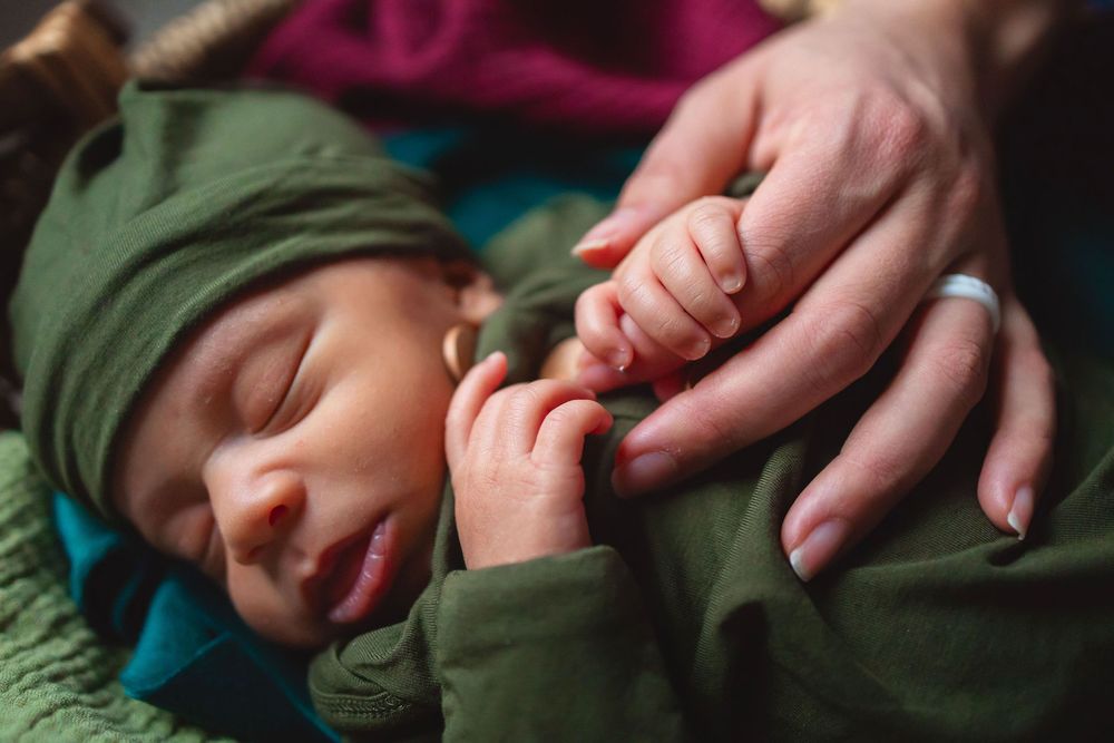Small sleeping newborn wearing green outfit grasps adult finger while resting on colorful blanket.