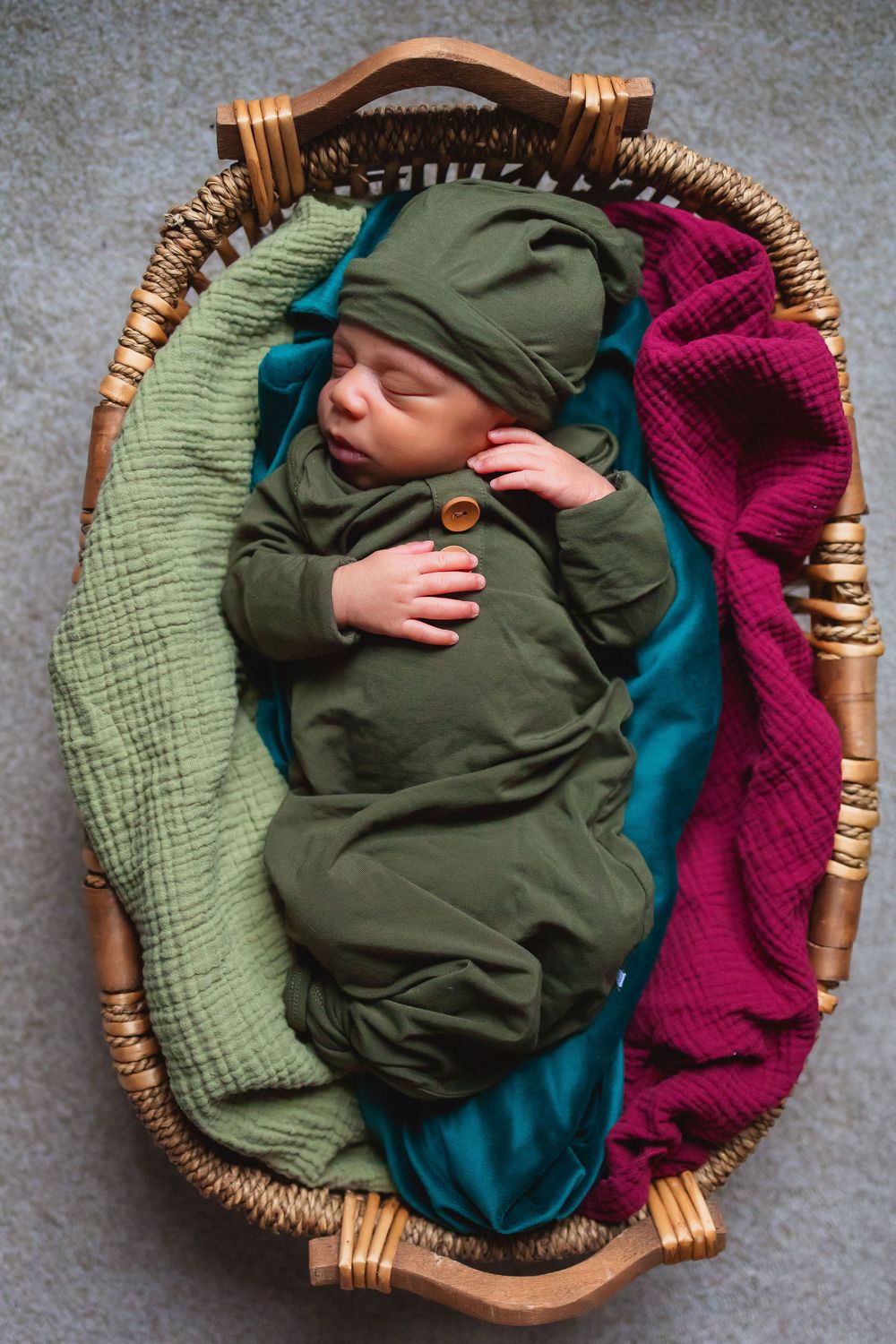 A sleeping infant swaddled in olive green lies in a woven basket with teal and burgundy textiles.