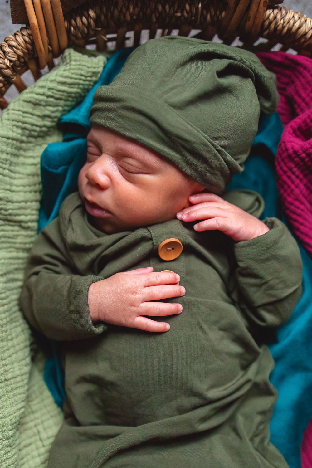 A sleeping baby wearing a cozy green outfit with matching hat rests on colorful knit blankets.