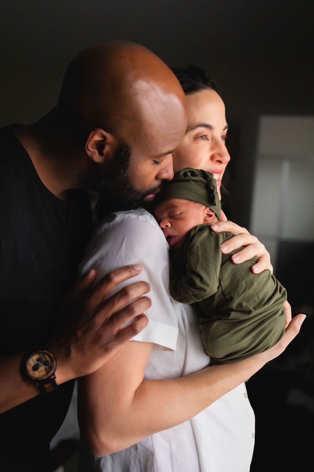 A tender family moment as parents embrace their sleeping newborn baby in dark, moody lighting.