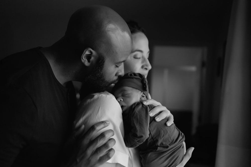 A tender black and white portrait of parents holding and gazing lovingly at their newborn baby in dim lighting.