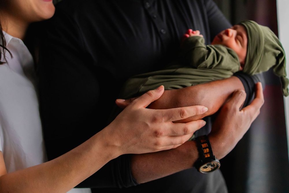 Close-up of gentle hands cradling and supporting a newborn baby wrapped in an olive green blanket.