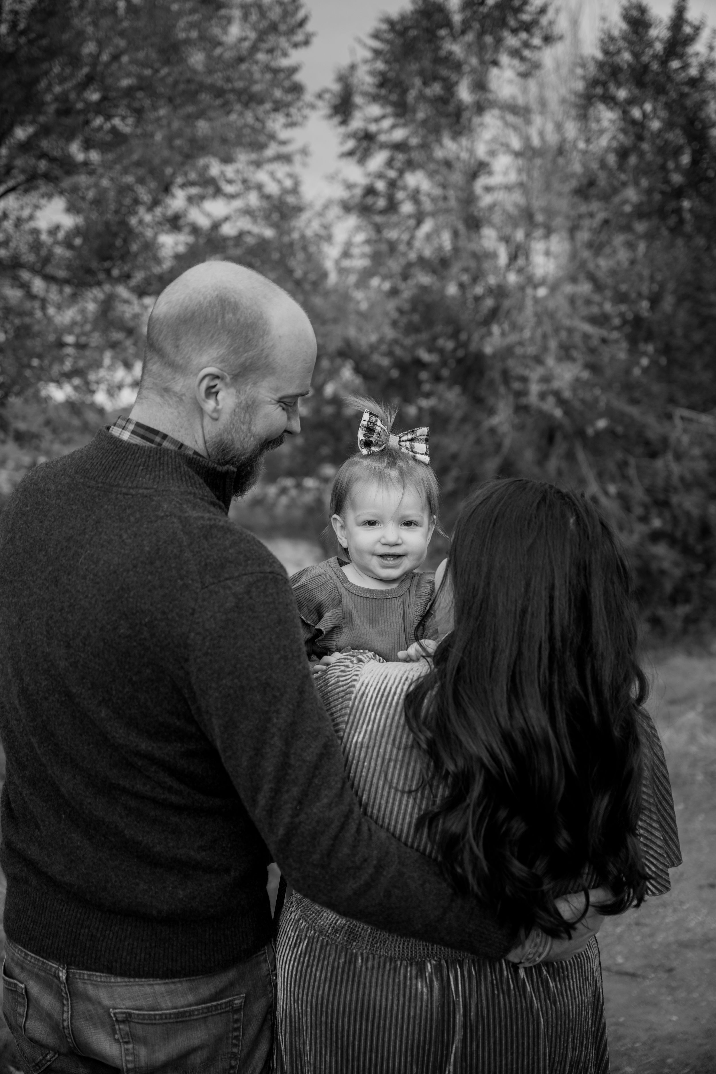 A black and white family portrait taken outdoors showing parents embracing their toddler against a wooded background.