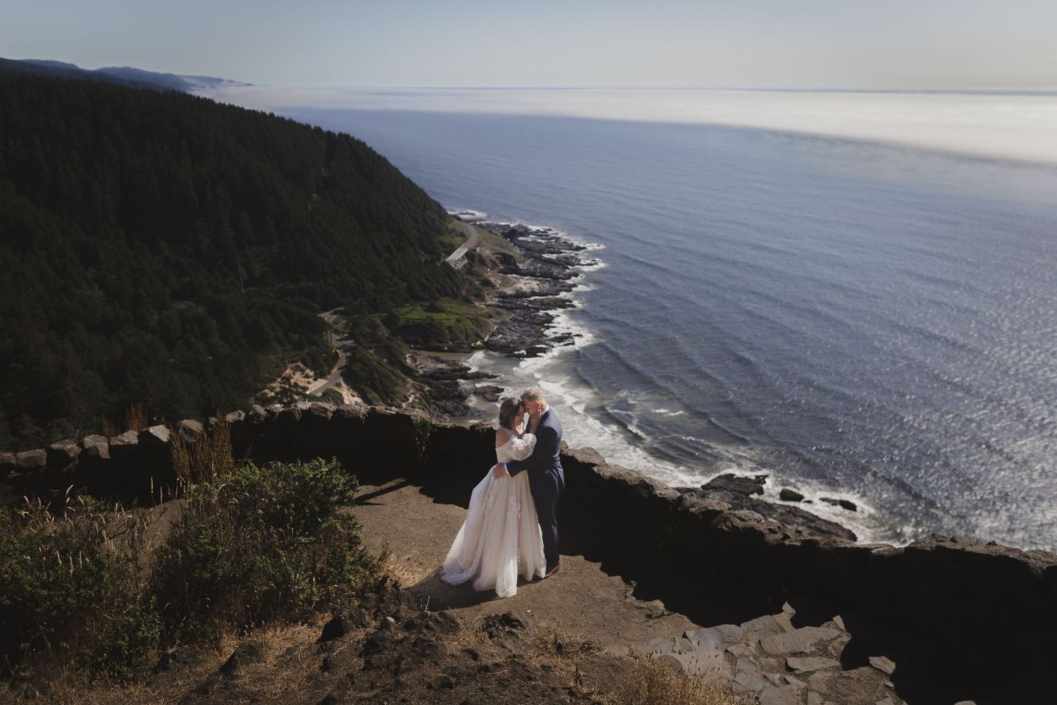 Scenic coastal wedding portrait overlooking rugged cliffs and crashing waves along dramatic Pacific Ocean shoreline.