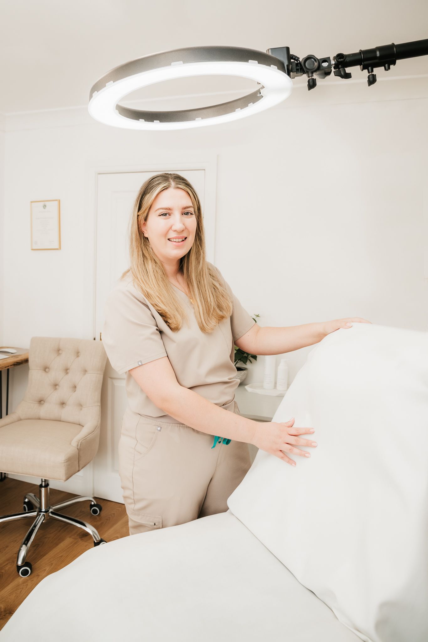 Healthcare professional in beige scrubs preparing a treatment bed under a ring light in a medical spa setting.