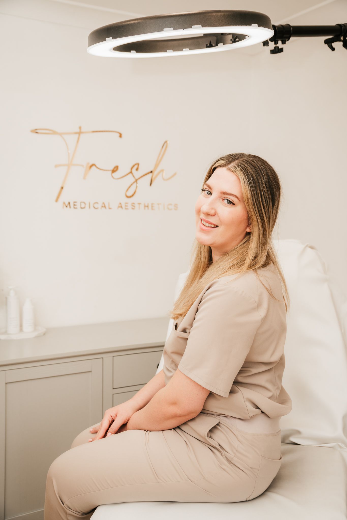 Medical aesthetician in beige scrubs sits on treatment chair under ring light at Fresh Medical Aesthetics clinic.