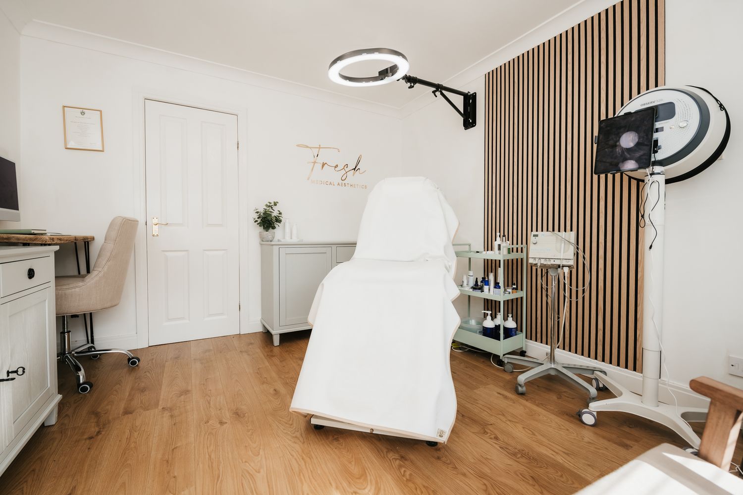 Modern beauty treatment room with wood slat wall, white treatment chair, and professional skincare equipment on rolling cart.