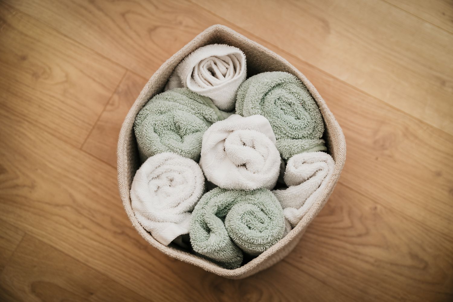 A woven basket filled with rolled sage green and white towels arranged in a spiral pattern on wooden flooring.