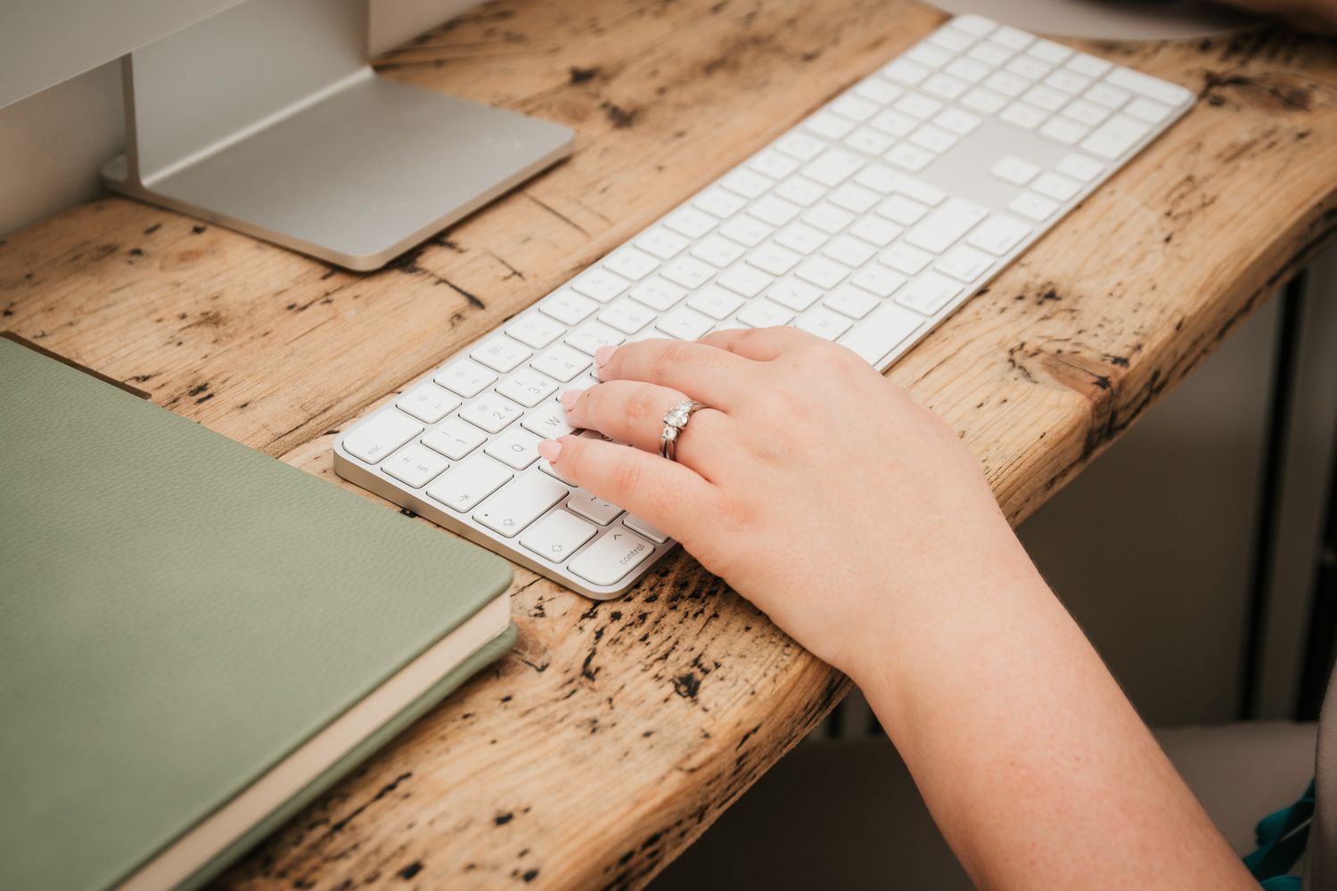 Hand wearing silver ring typing on white wireless keyboard on rustic wooden desk surface.