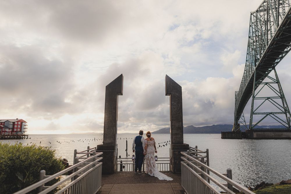 Silhouettes stand on a scenic dock overlooking the Astoria-Megler Bridge spanning the Columbia River at sunset.