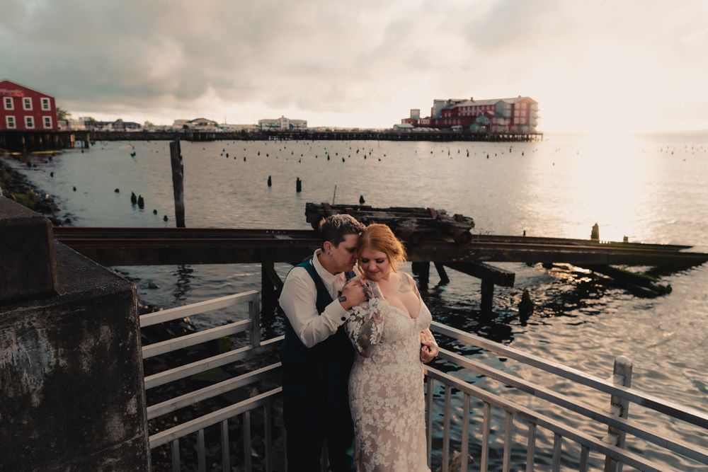 Newlyweds embrace on stone steps near a harbor at sunset with wooden pilings visible in the water.