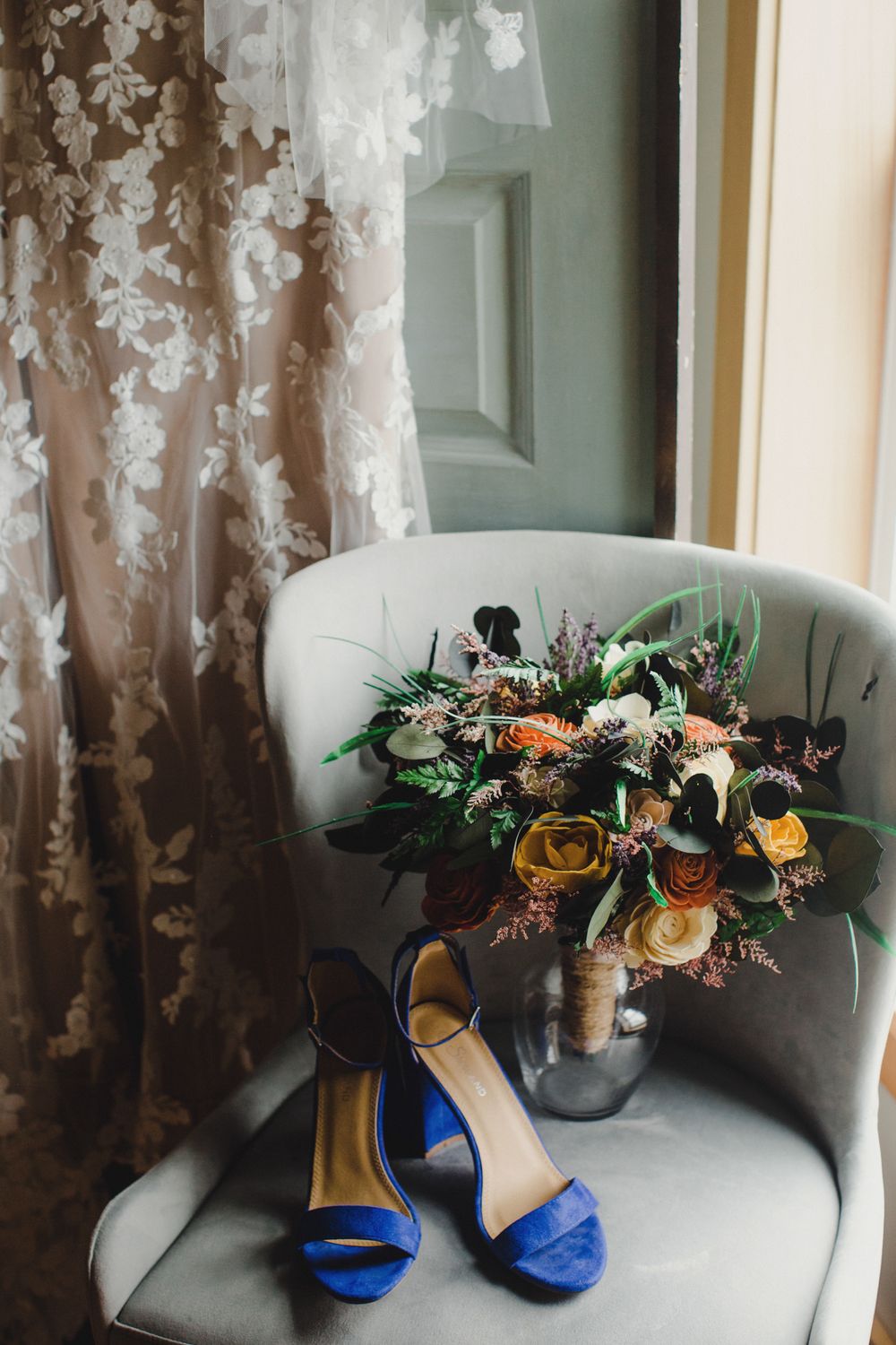 Wedding bouquet with orange and yellow roses sits on gray armchair beside bright blue bridal shoes.