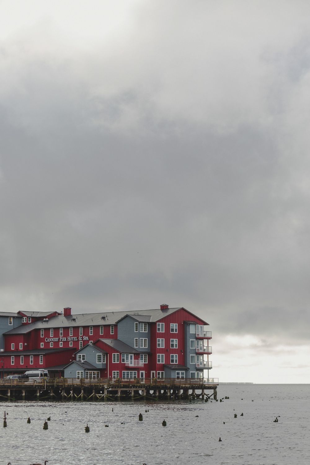 A large red and gray waterfront hotel building stands against a moody cloudy sky above a rocky beach at low tide.