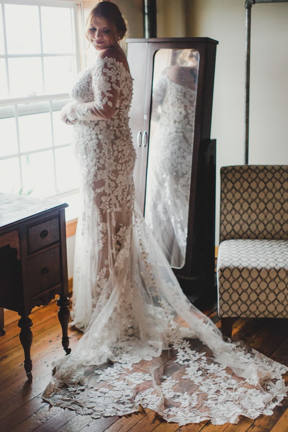A wedding dress with intricate lace details and long train is displayed in a sunlit room with vintage furniture.