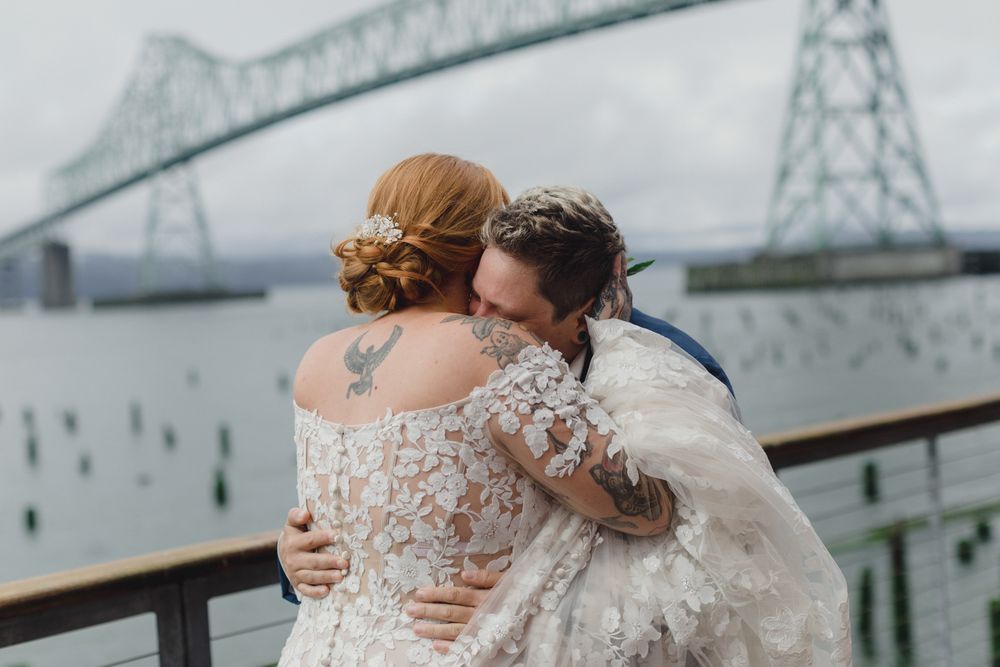 Couple embraces during wedding photoshoot with Astoria-Megler Bridge in background on overcast day.