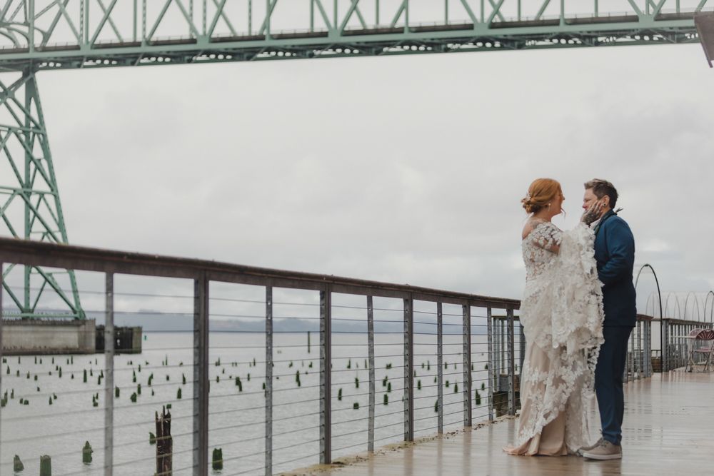 Two people stand on a waterfront boardwalk beneath a large steel bridge on an overcast day.