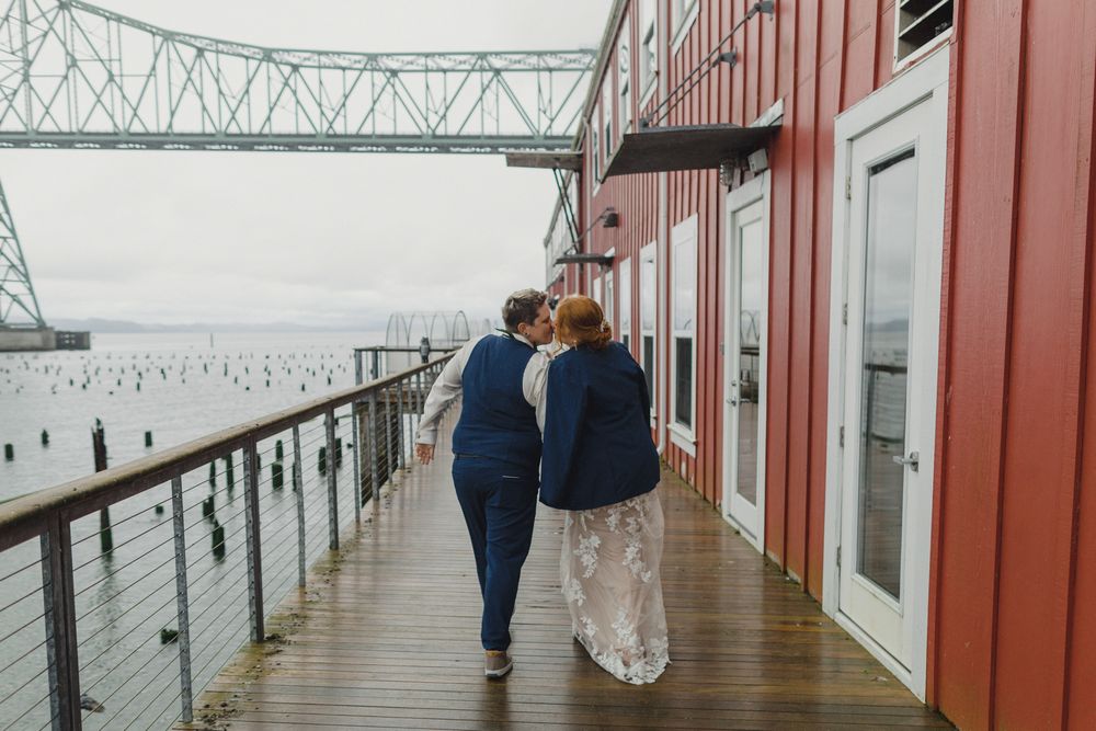 A couple walks together on a waterfront boardwalk with industrial bridge in the background.