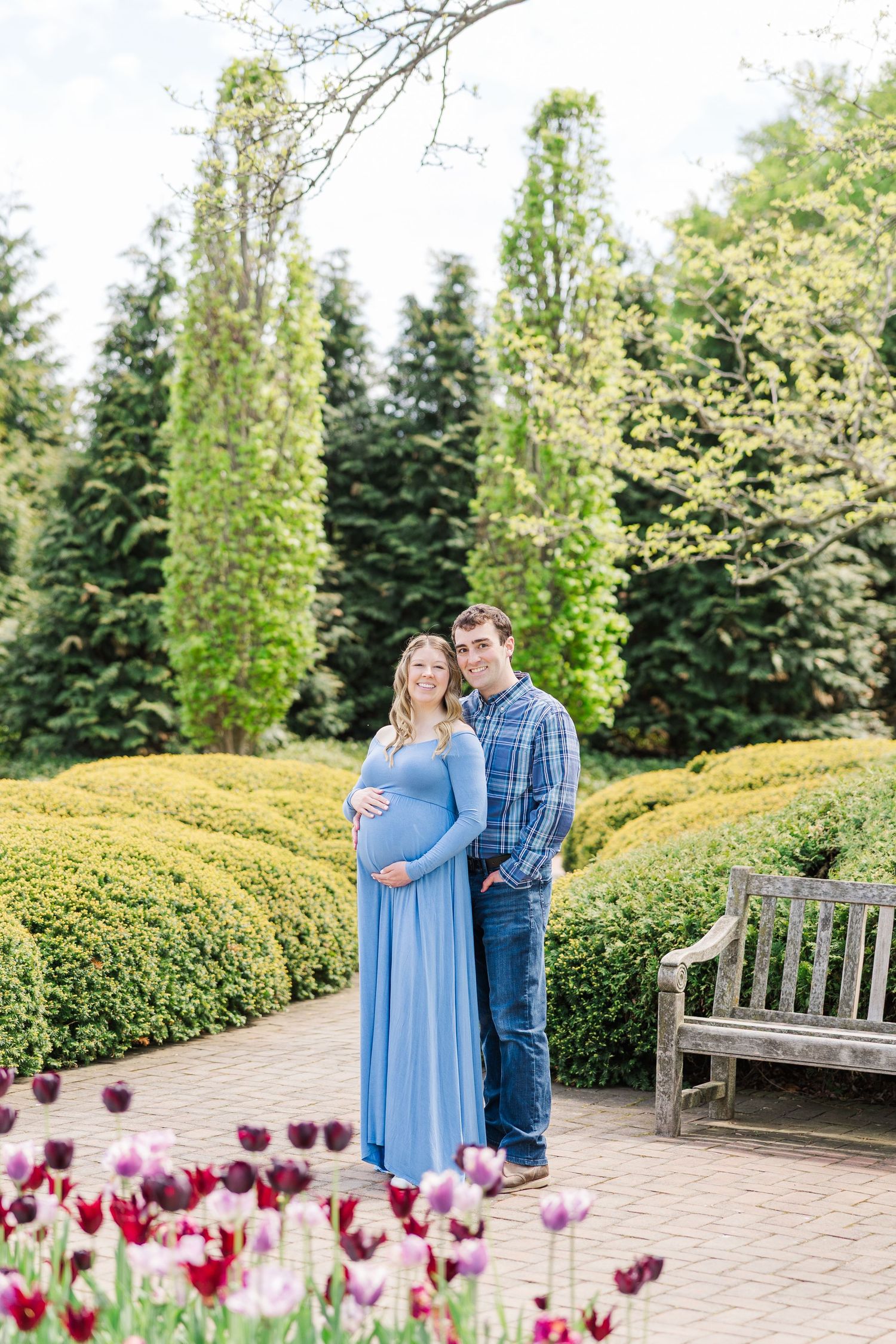 A couple stands together on a garden path lined with tulips and tall evergreen trees near a wooden bench at Chicago Botanic Garden.