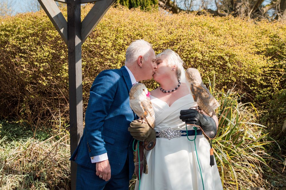 A couple shares a tender moment under a wooden beam on their wedding day in a sunny outdoor garden setting.