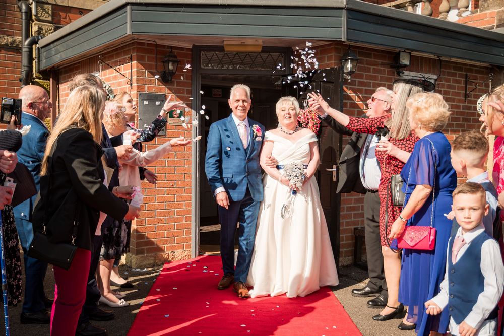 Wedding party exits building onto red carpet as guests toss confetti in celebration.