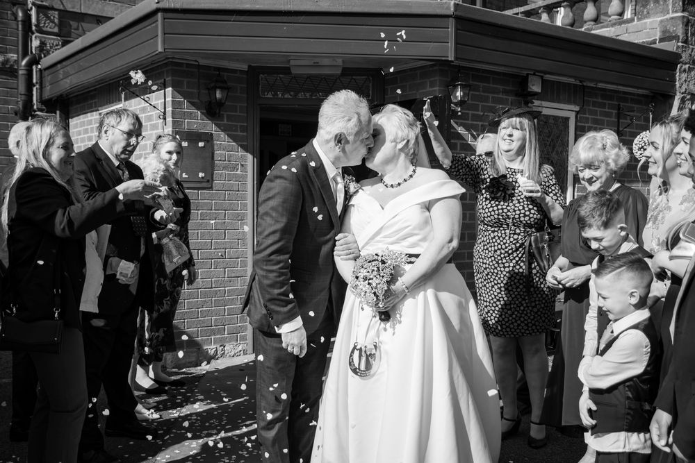 Black and white photo of a wedding couple kissing while guests throw confetti outside a brick building.