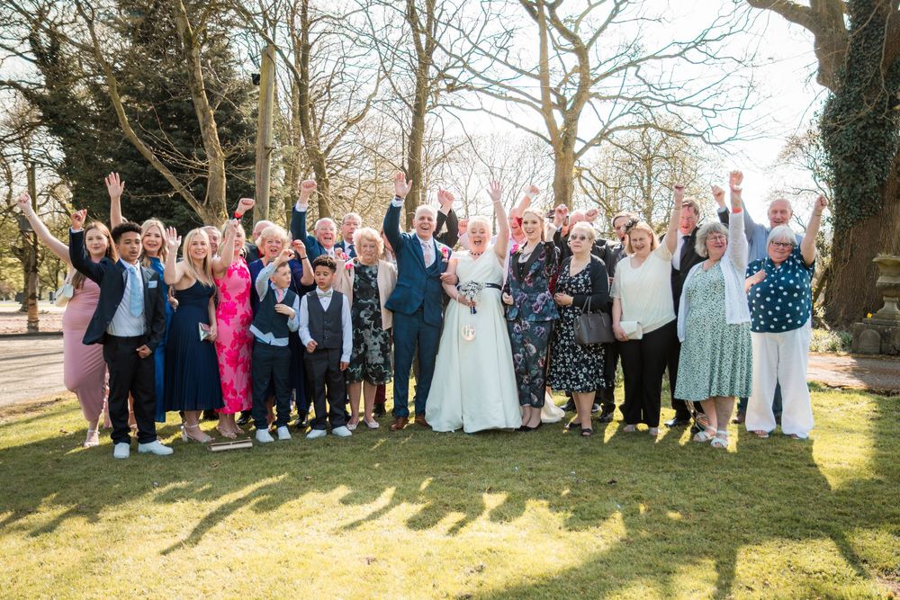 Wedding party and guests celebrate outdoors on a sunny day with trees casting long shadows on grass.