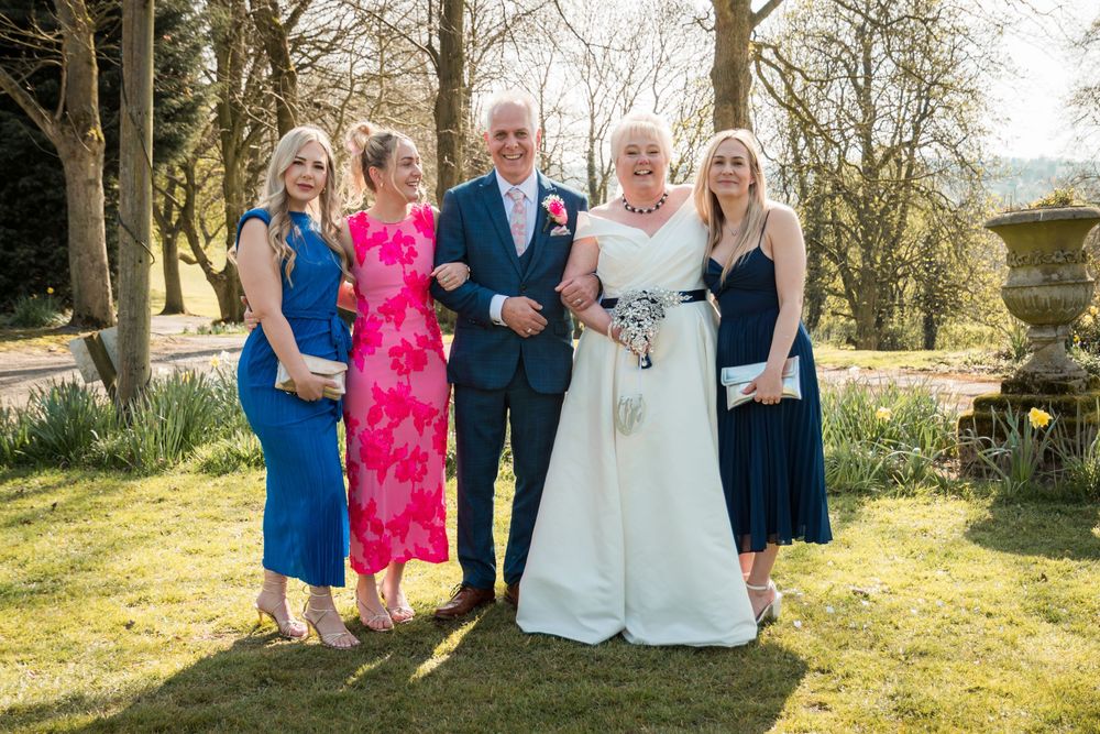Group in formal attire poses together on sunny lawn with trees in background during outdoor celebration.