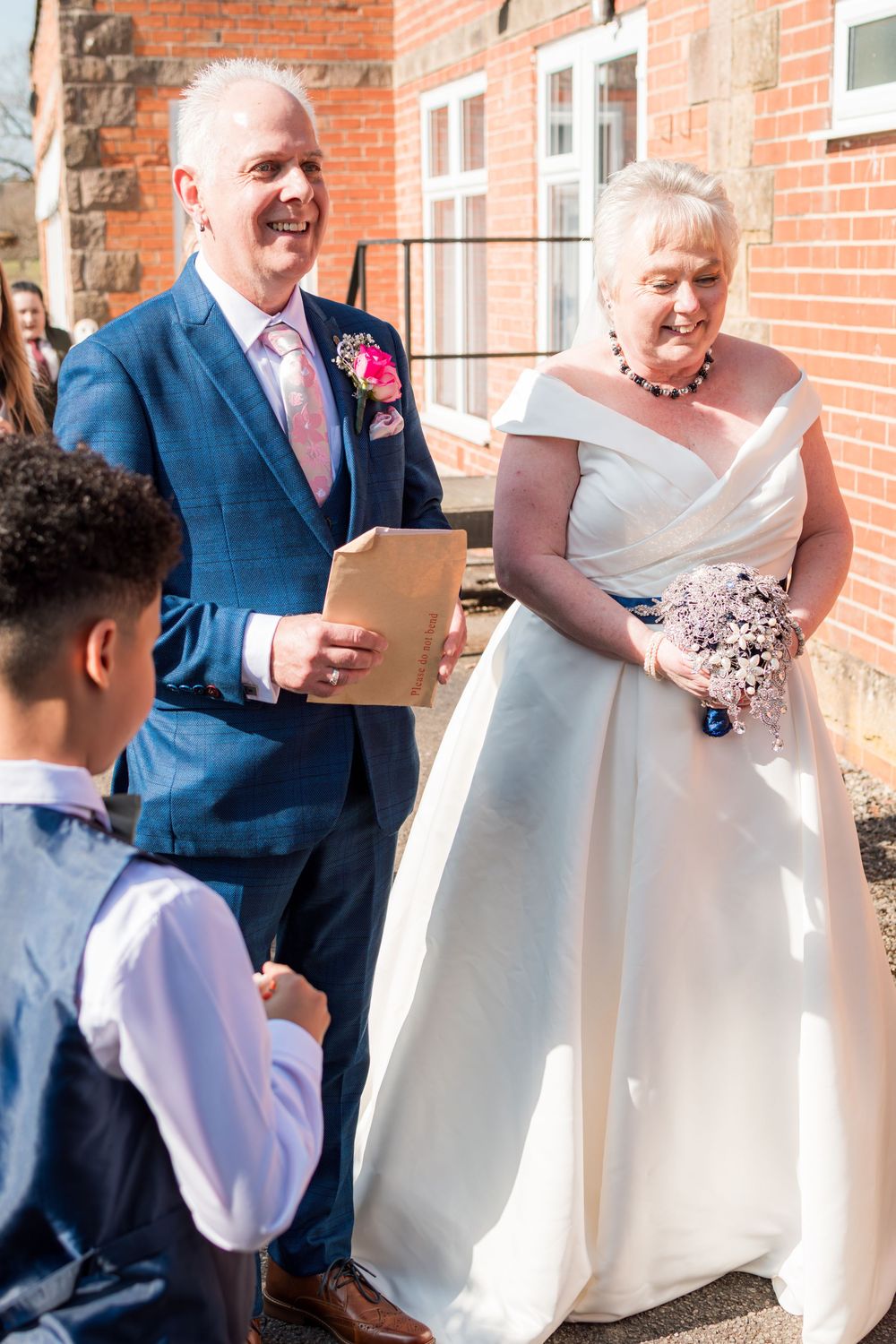 A sunny wedding ceremony outdoors with the couple and a young attendant standing by a brick building.