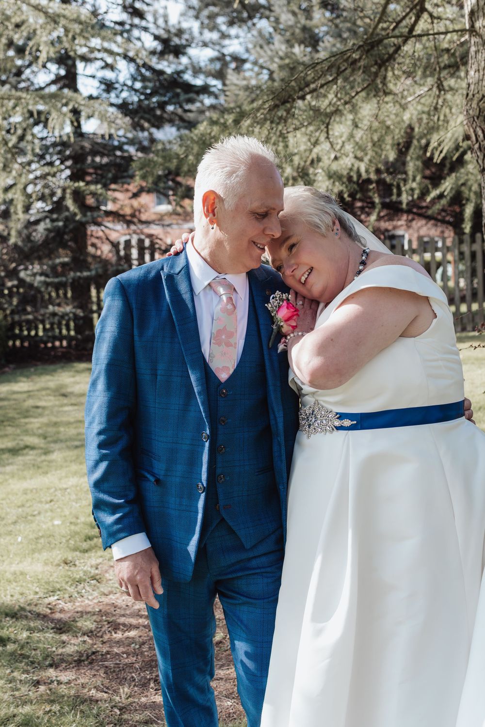 Intimate wedding portrait of newlyweds in a garden setting with blue suit and white dress with navy sash detail.