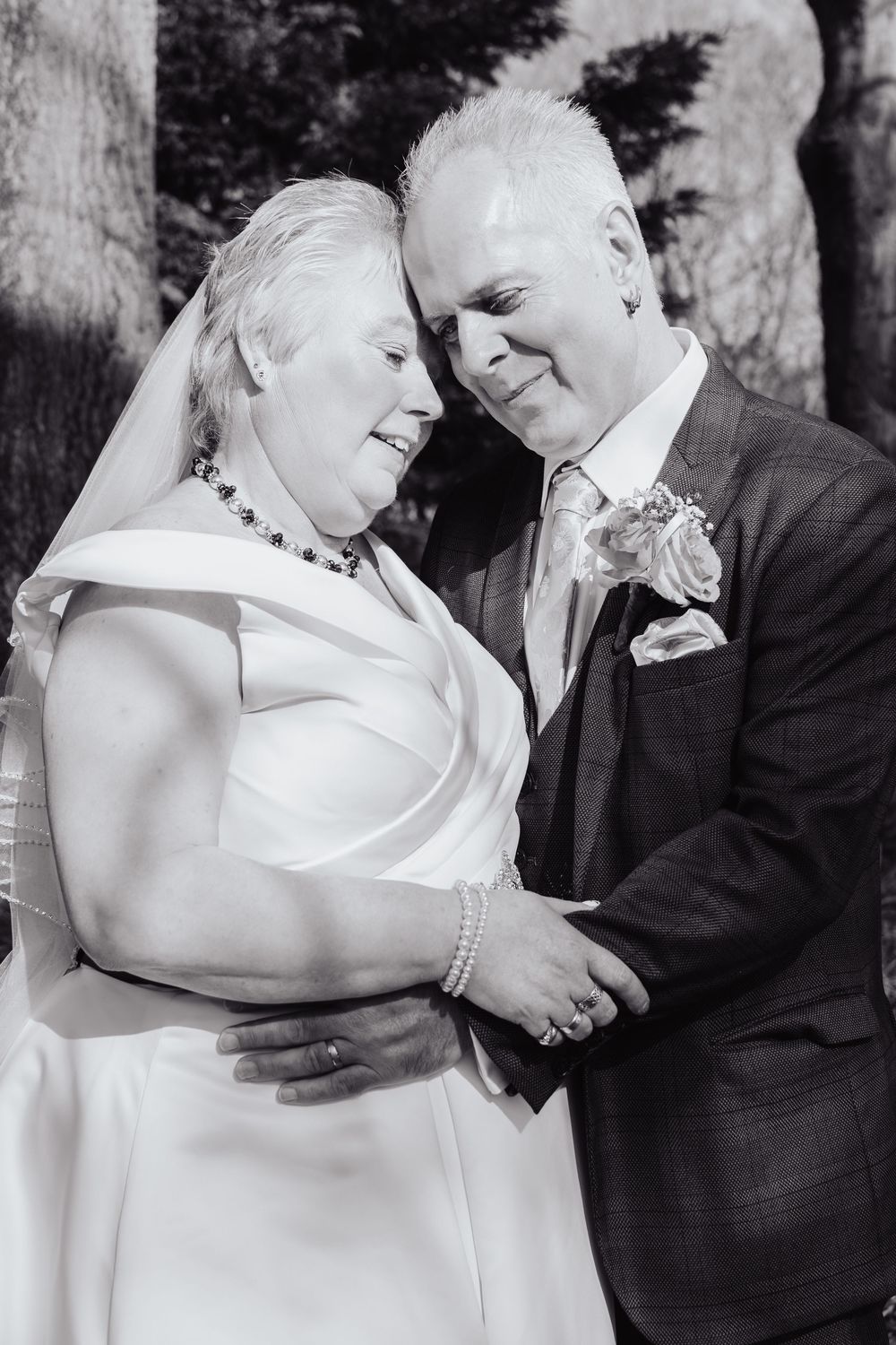 Black and white wedding photograph showing a tender moment between newlyweds in formal attire embracing outdoors.