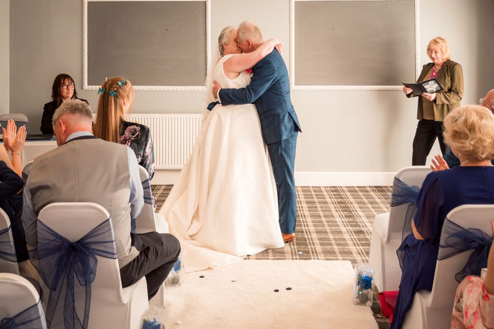 A couple embraces during an indoor wedding ceremony while guests look on from white covered chairs in an elegant venue.