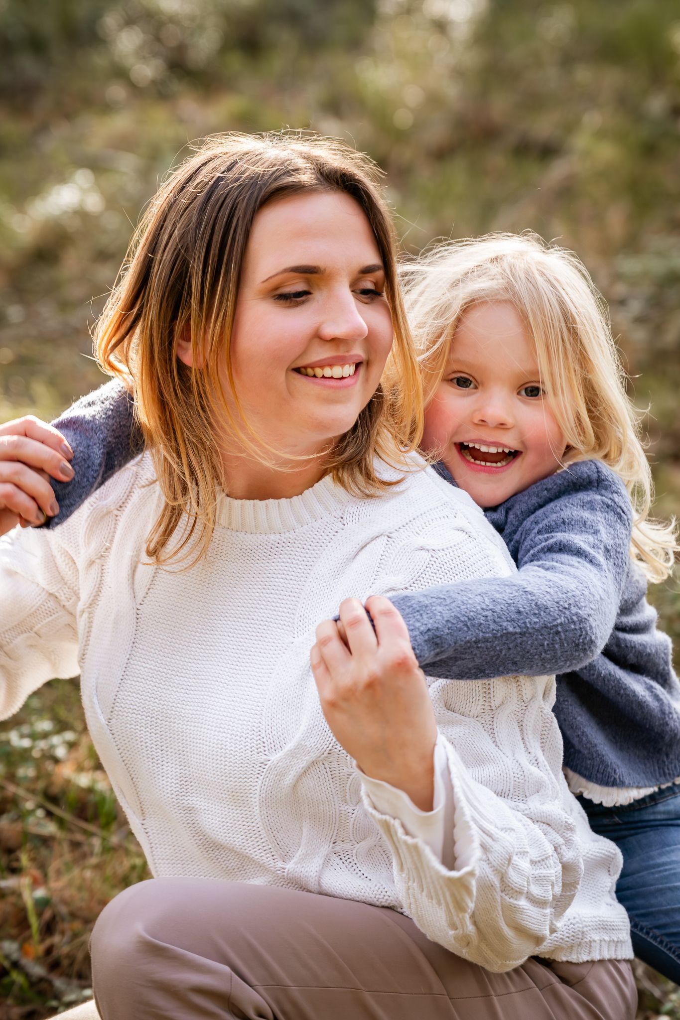 Mamma sitter på huk i skogen med en glad dotter som kramar om henne bakifrån. Fotograf, familjefotograf, Umeå, Västerbotten