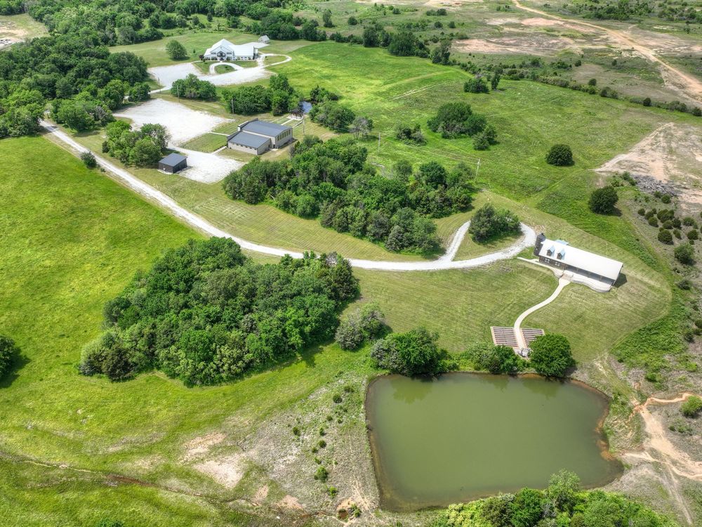 Aerial view of rural property with winding driveway, scattered buildings, green pastures and small pond in countryside setting.