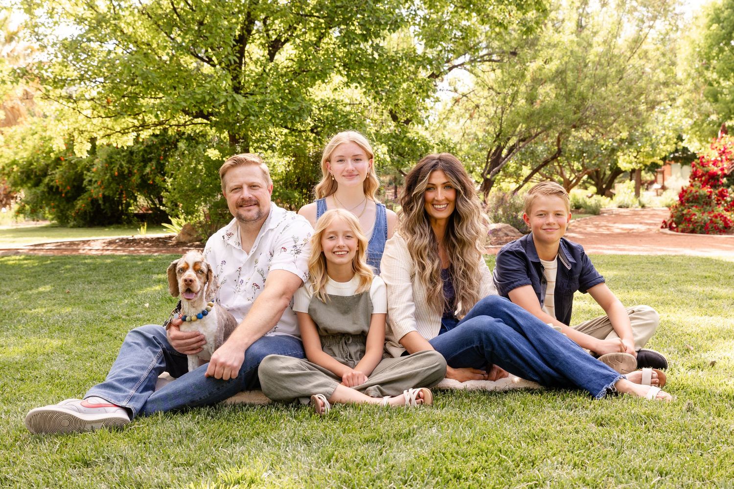 mom dad 3 kids and a dog sitting on a blanket on a green field with a shade tree on the background