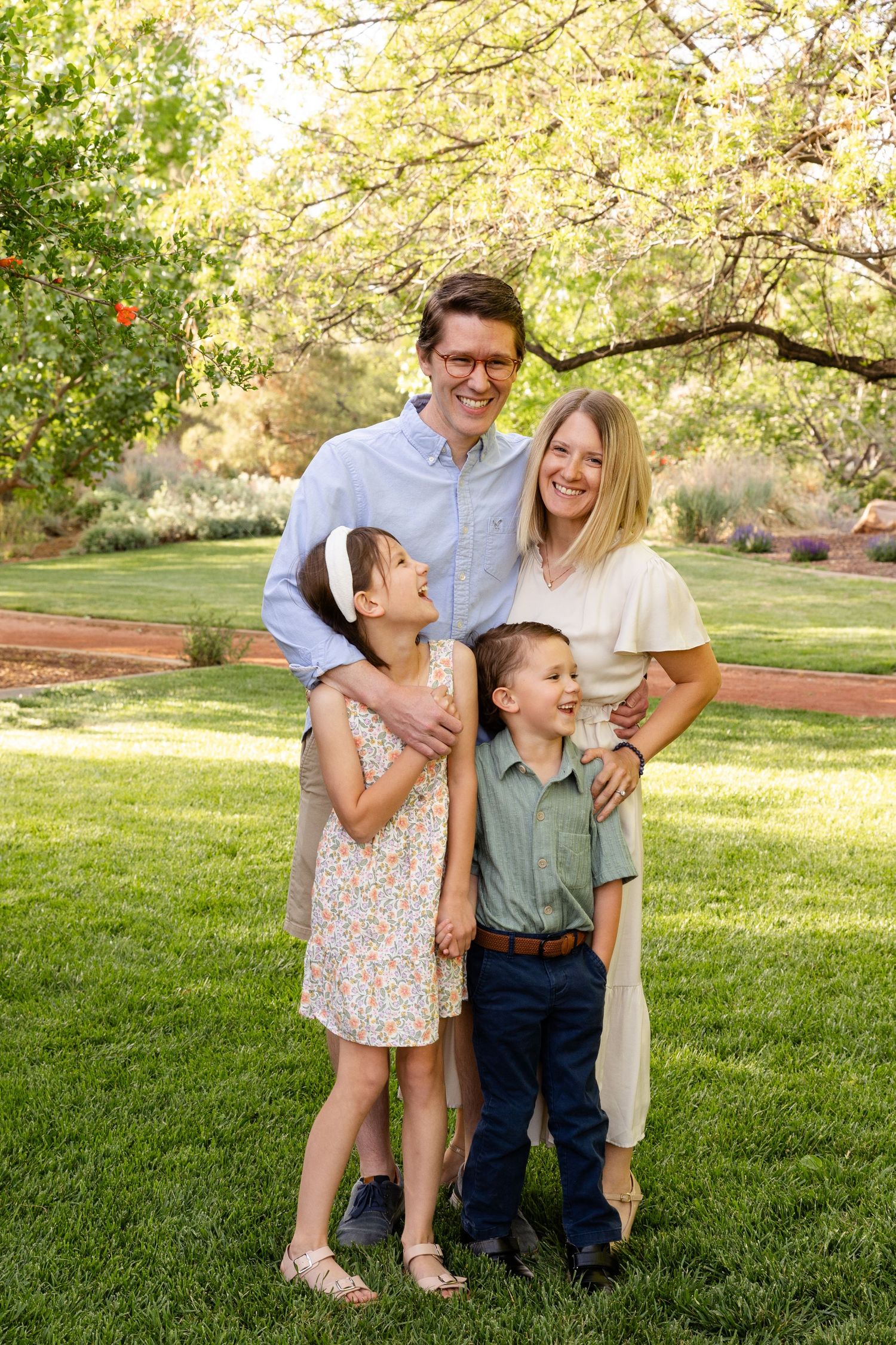 mom dad and 2 young children standing on a grassy field with a big shade tree above them. laughing