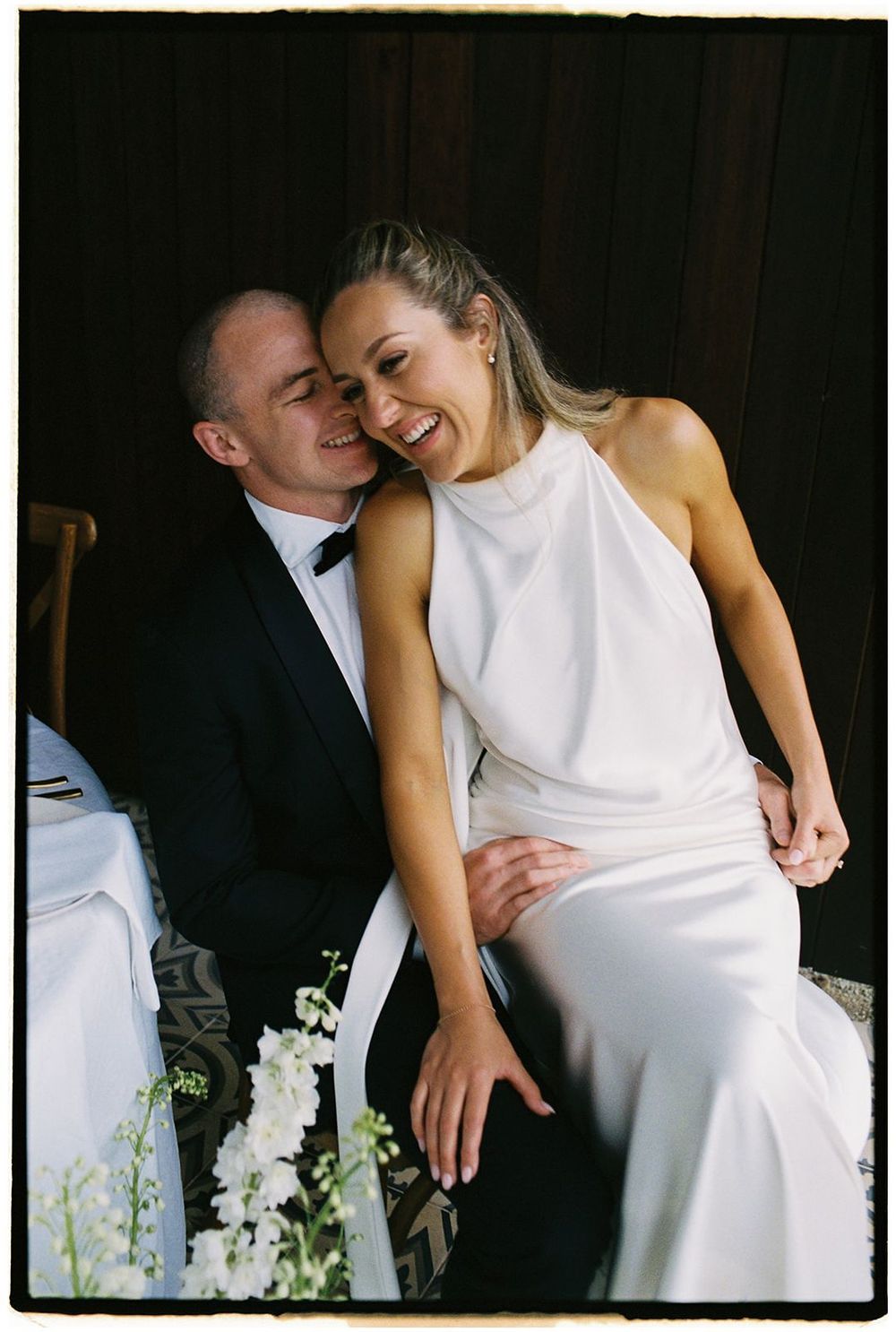 A couple shares a joyful candid moment in formal wedding attire with white orchids in the foreground.