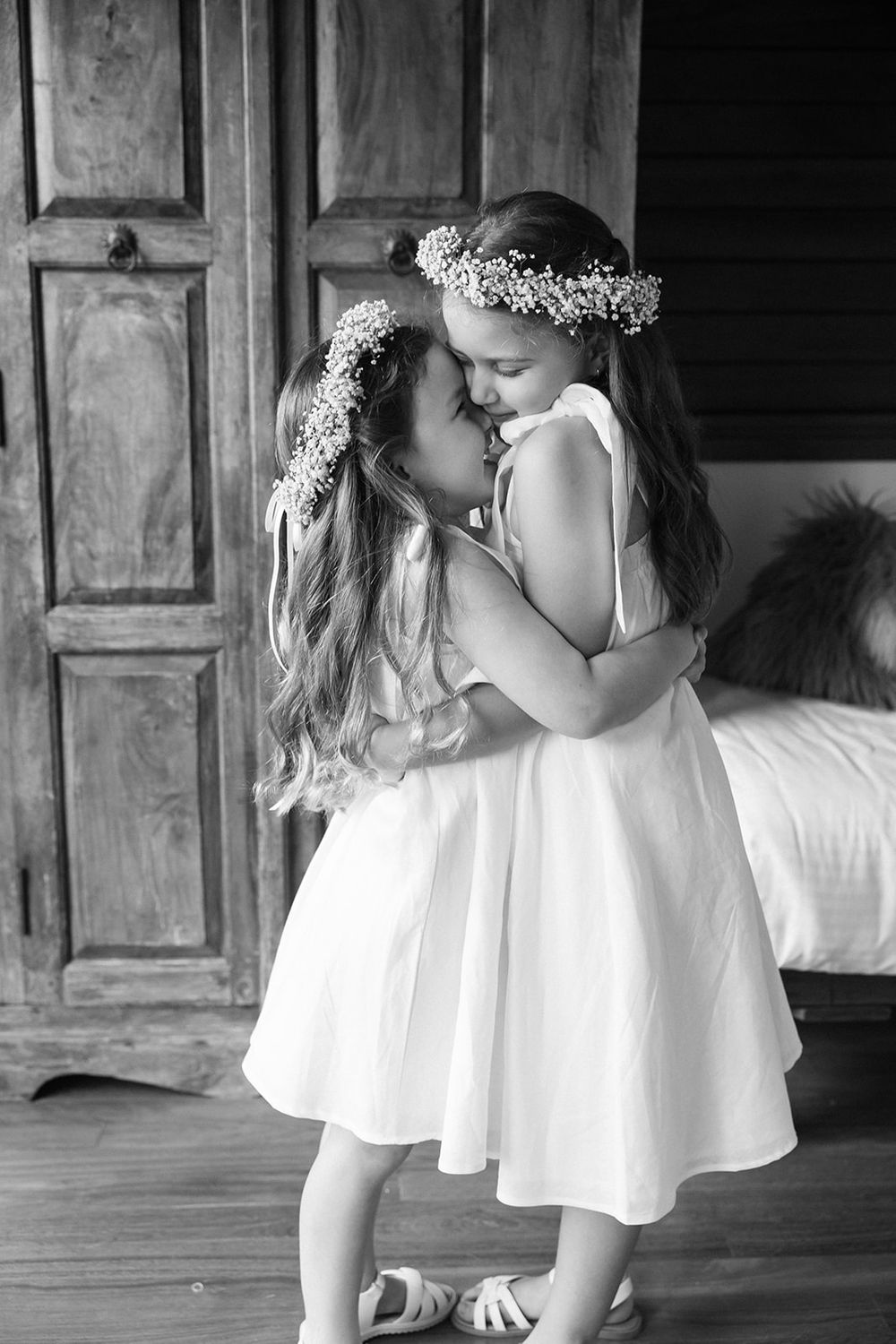Two girls in white dresses and floral crowns share a tender moment together during a wedding celebration.