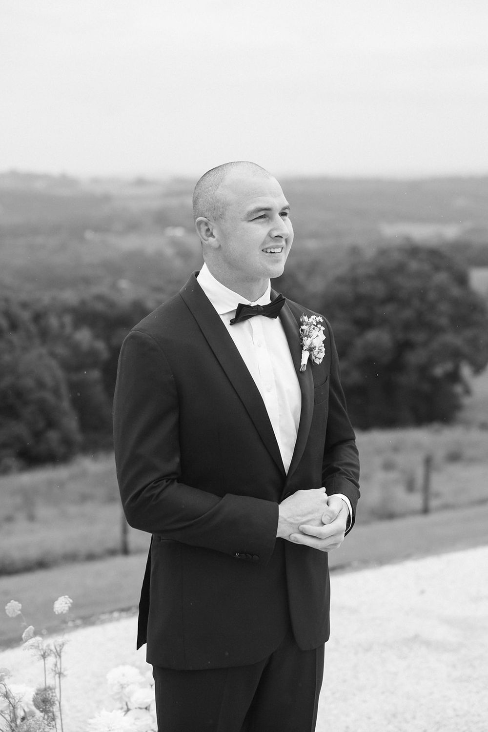Groom in classic black tuxedo smiles during outdoor wedding ceremony against scenic hillside backdrop.
