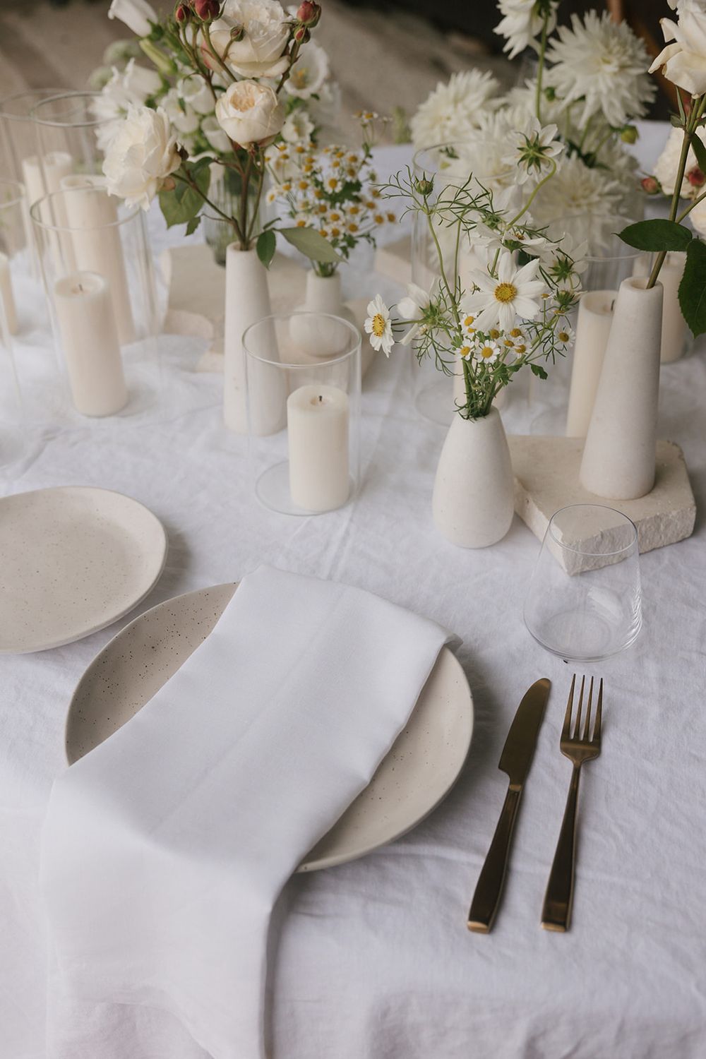 White place setting with floral arrangements and glass candle holders on a linen tablecloth.