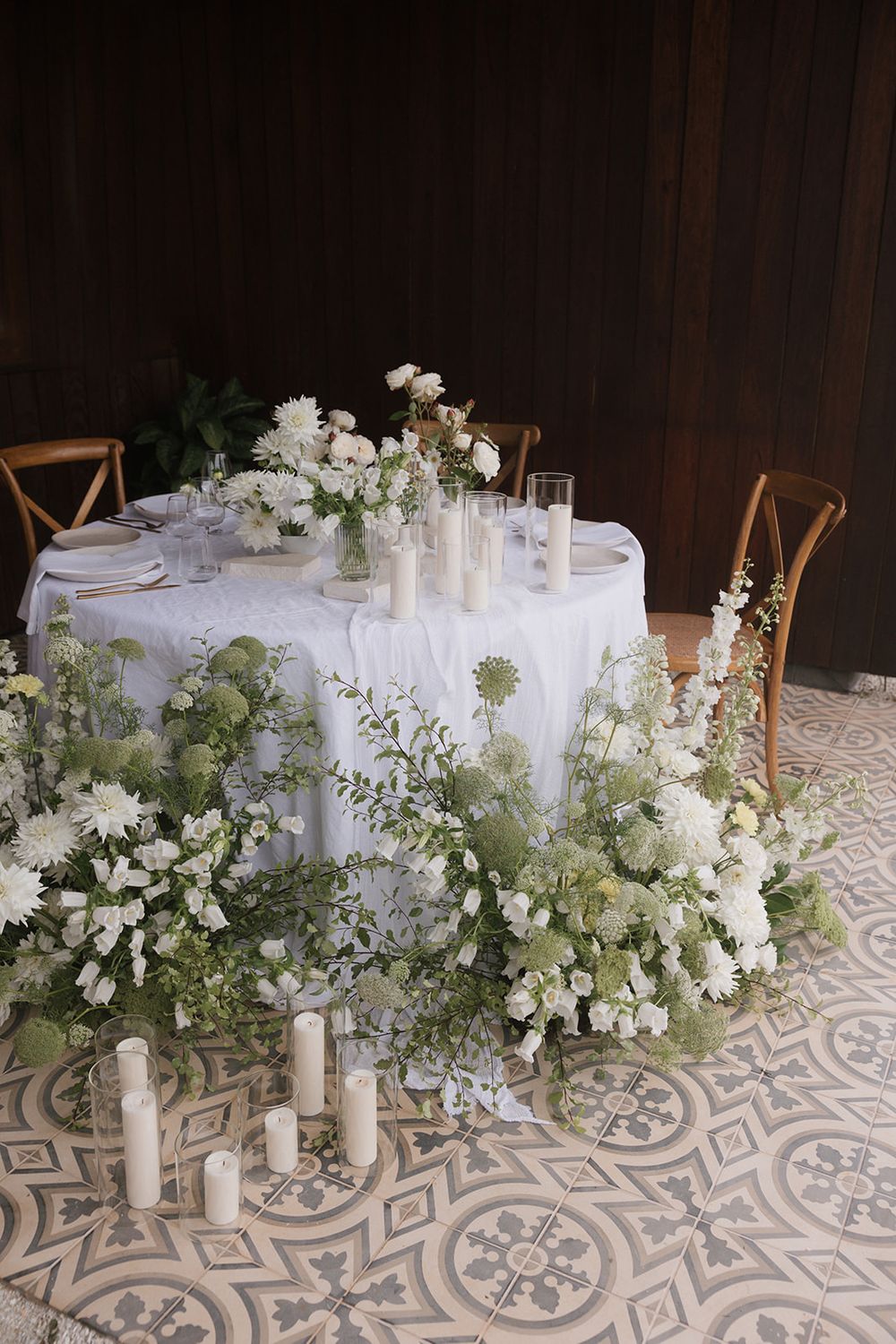 Wedding table decor featuring white flowers, candles and wooden chairs on a patterned floor.