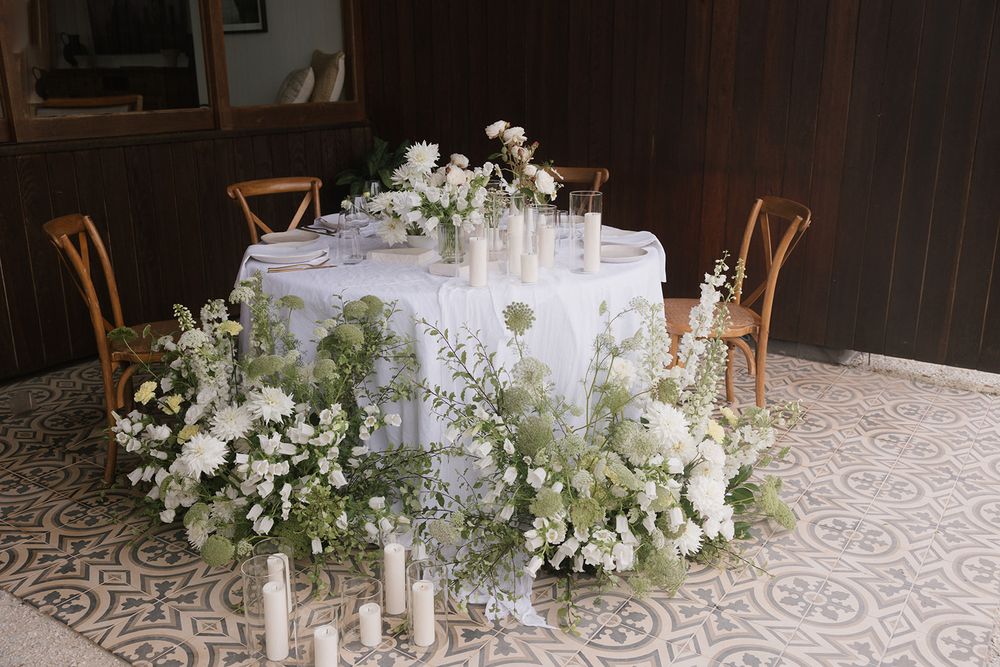 Round wedding table with lush white floral arrangements and taper candles on patterned floor.