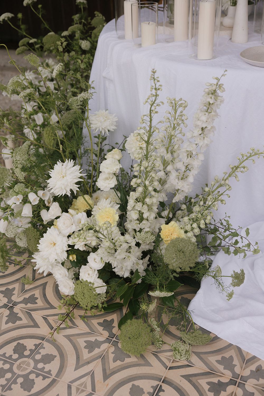 White floral arrangement with delphinium and candles on a patterned tile floor.