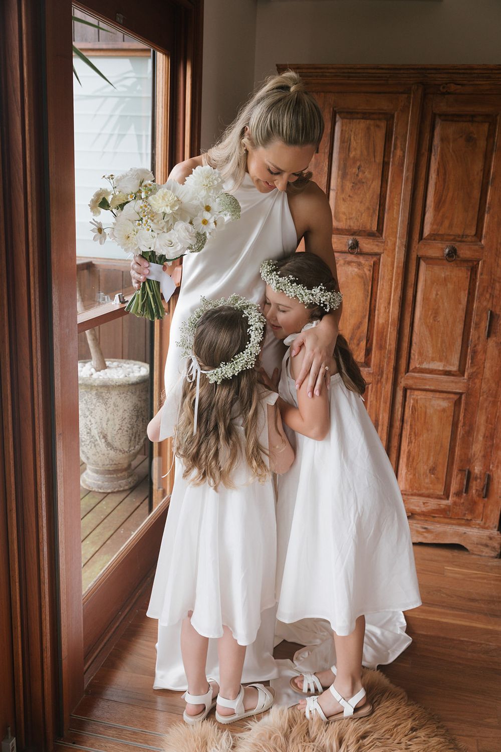 Wedding preparation scene in white dresses with floral crowns by wooden wardrobe.