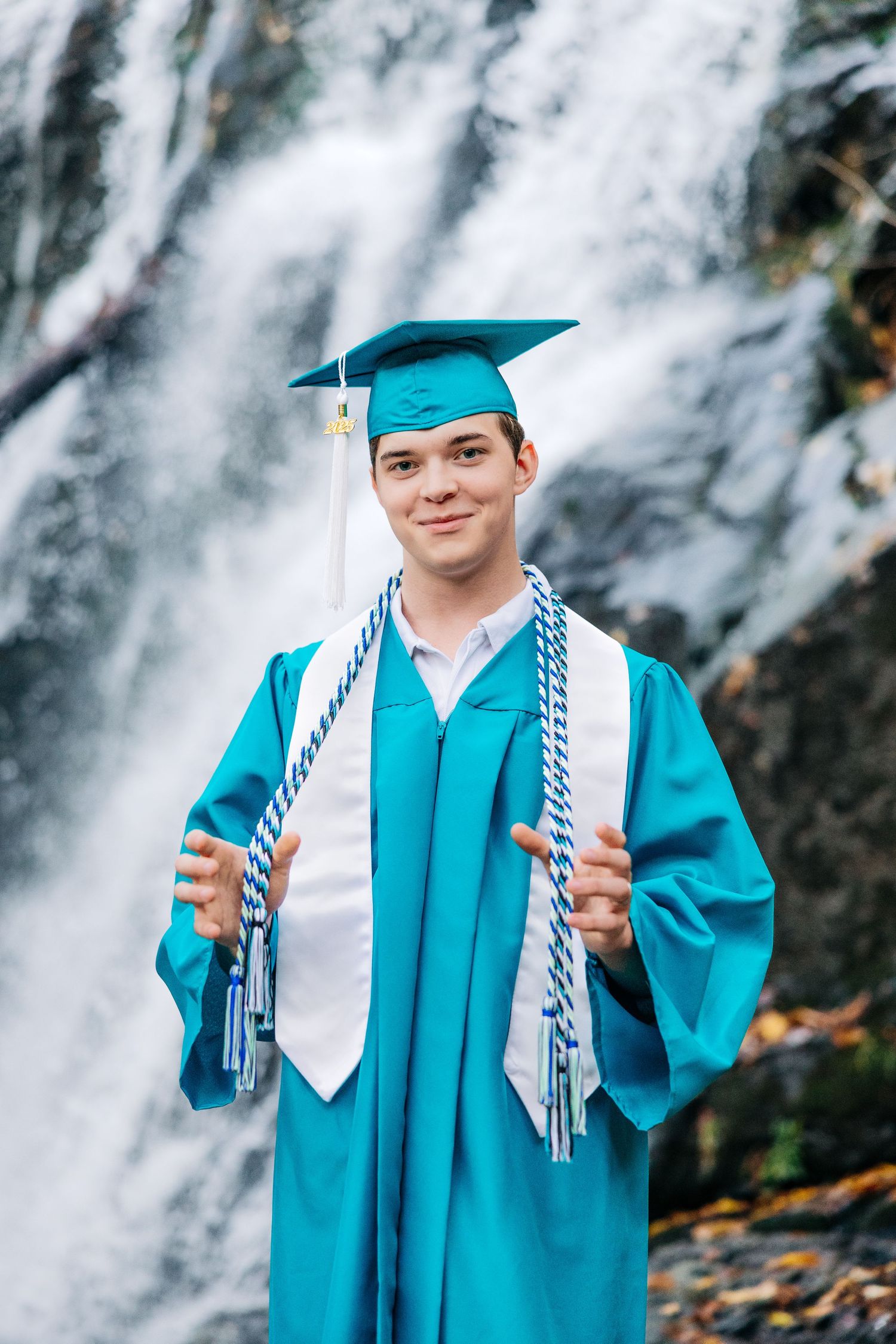 Graduate in turquoise cap and gown holding white and silver honor cords stands in front of cascading waterfall.