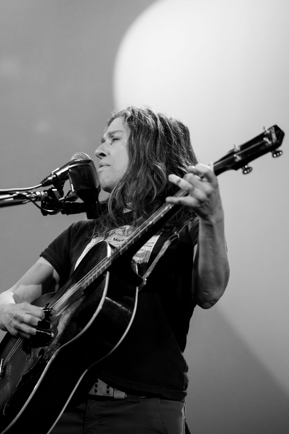 A musician in black performs passionately at a microphone while playing an acoustic guitar in black and white.