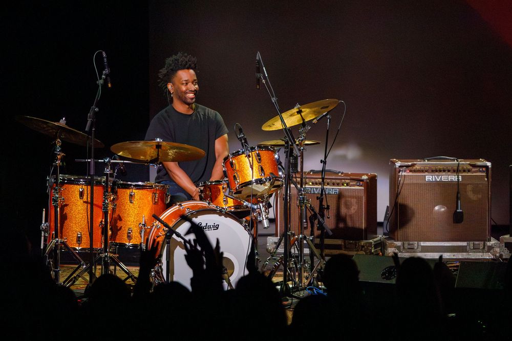 Drummer playing orange drum kit on dark stage with amplifiers in background.