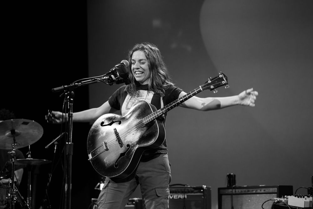 A guitarist performs energetically on stage with arms outstretched while singing into a microphone in monochrome.
